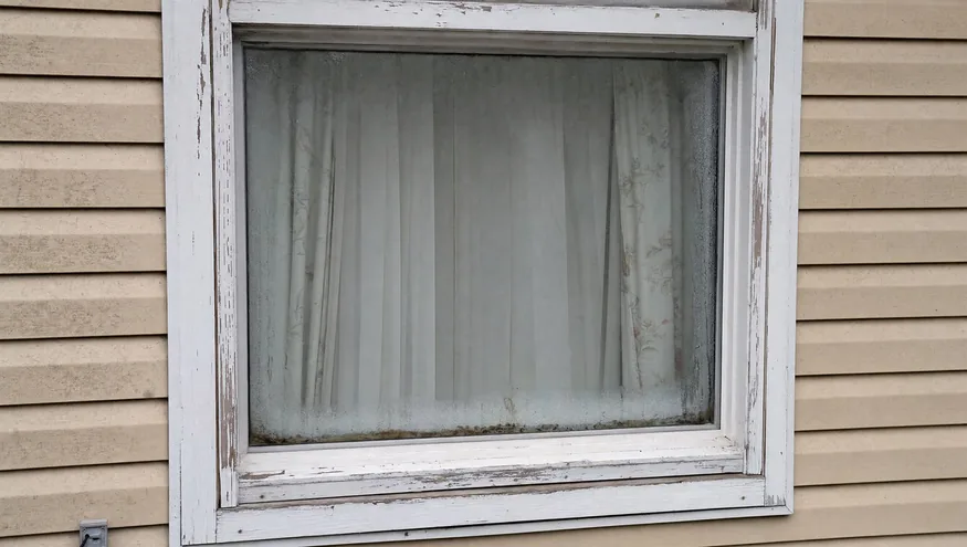An outdated, drafty single-pane wood-framed window with peeling paint and condensation damage
